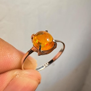 Puede incluir: Anillo de plata con una piedra de ópalo ovalada. El ópalo muestra un color naranja cálido con destellos rojos y verdes. La banda del anillo es de plata pulida, resaltando el color y brillo de la piedra preciosa. Una joya.