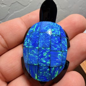 Puede incluir: Una figura de tortuga artesanal con un caparazón de mosaico de piedras iridiscentes azules y verdes. La cabeza, las aletas y la cola son negras. La figura se sostiene en la mano de una persona.