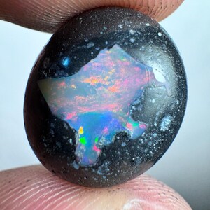 Puede incluir: Una piedra preciosa de ópalo negro pulido, de forma ovalada. El ópalo presenta un vibrante juego de colores, con destellos de azul, verde, rosa y naranja. La piedra se sostiene entre dos dedos, mostrando su tamaño y detalle.