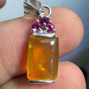 Puede incluir: Un colgante de plata con una piedra de ópalo naranja rectangular y una corona de piedras rosas.