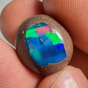 Puede incluir: Una piedra preciosa de ópalo pulida, de forma ovalada. El ópalo presenta una matriz marrón con una exhibición vibrante de color, incluyendo tonos azules, verdes y rosas. La piedra preciosa se sostiene entre los dedos.