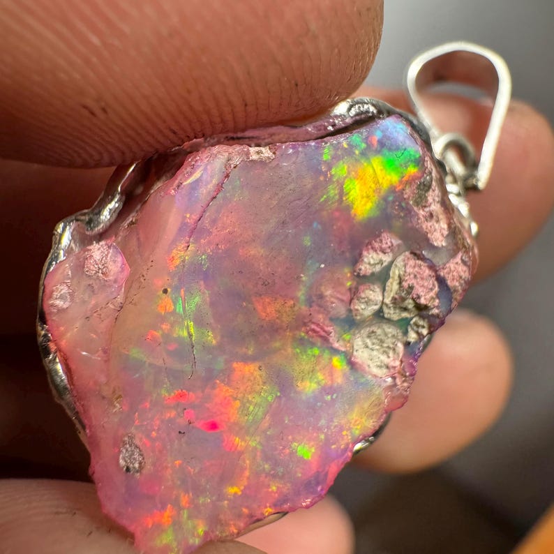 May include: A pink opal pendant with a silver setting. The opal has a rough, natural shape and displays a rainbow of colors.