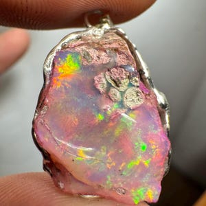 May include: A pink opal pendant with a silver setting. The opal has a rough, natural shape and displays a rainbow of colors, including pink, green, and orange.