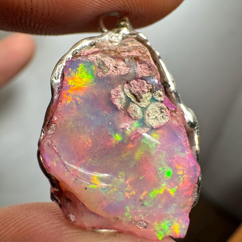 May include: A pink opal pendant with a silver setting. The opal has a rough, natural shape and displays a rainbow of colors, including pink, green, and orange.