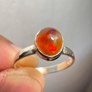 May include: A silver ring featuring an oval-shaped orange opal gemstone. The opal displays a red and purple flash.