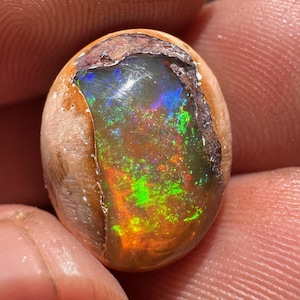 May include: A polished oval opal gemstone with a rough, brown exterior. The opal's interior displays vibrant colors, including green, blue, and orange, creating a play-of-color effect. The gemstone is held between fingers.