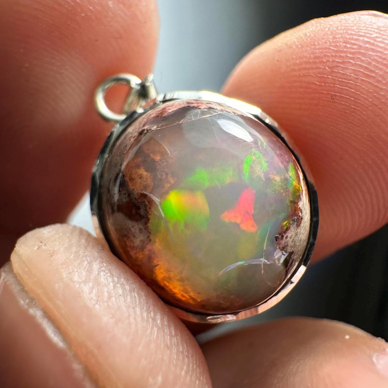 May include: A silver pendant with a round, iridescent opal stone. The opal has a brown, green, and red play of color.