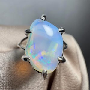 May include: A silver ring featuring a large, oval-shaped opal gemstone. The opal is a milky white colour with flashes of blue, green, and pink.