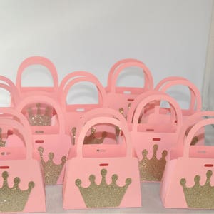 May include: A collection of pink paper purses with arched handles. Each purse features a gold glitter crown on the front. The purses are designed for party favors or gifts, with a princess or royal theme.