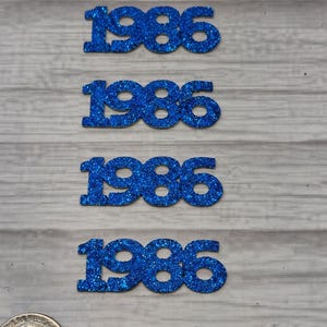 May include: Several blue glitter-covered number cutouts of "1986" are arranged on a light gray wooden surface. A U.S. quarter is included for size comparison. The numbers are evenly spaced and have a sparkly texture.