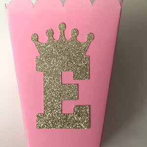 May include: Pink cardboard favor box with a scalloped edge. The box has a gold glitter crown and the letter "E" on the front.