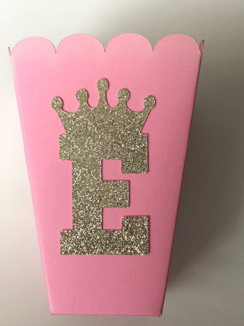 May include: Pink cardboard favor box with a scalloped edge. The box has a gold glitter crown and the letter "E" on the front.