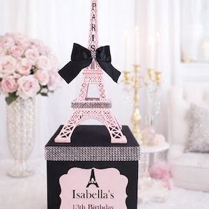 May include: A pink Eiffel Tower ornament, with the word "PARIS" on it, sits on a black box. The tower has a black bow with a rhinestone detail. The box has a pink label that says "Isabella's 13th Birthday".