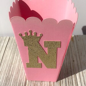 May include: Pink favor box with a scalloped edge. The box has a gold glitter crown and the letter N on the front.