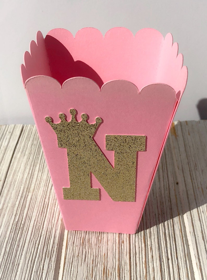 May include: Pink favor box with a scalloped edge. The box has a gold glitter crown and the letter N on the front.