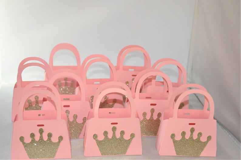 May include: A collection of pink paper purses with arched handles. Each purse features a gold glitter crown on the front. The purses are designed for party favors or gifts, with a princess or royal theme.