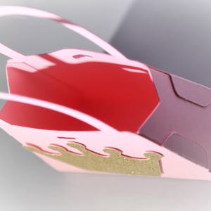 May include: A pink paper gift bag with a gold glitter crown design. The bag has a red interior and a handle. The bag is folded and has a shadow.