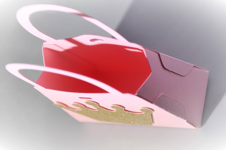 May include: A pink paper gift bag with a gold glitter crown design. The bag has a red interior and a handle. The bag is folded and has a shadow.