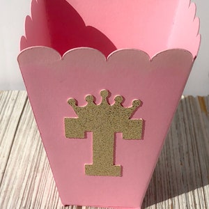 May include: Pink popcorn box with a scalloped edge and a gold glitter letter "T" with a crown on top.