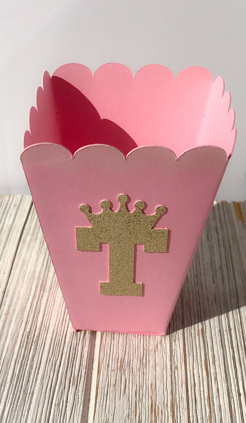 May include: Pink popcorn box with a scalloped edge and a gold glitter letter "T" with a crown on top.