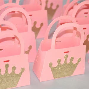 May include: Pink paper purse-shaped gift boxes with gold glitter crown designs. Each box has a handle and a small red accent. The boxes are arranged in a group, suggesting a party or event theme.