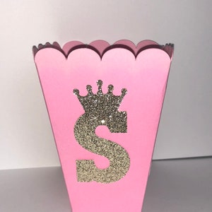 May include: Pink favor box with a scalloped edge. The box has a gold glitter crown and the letter "S" on the front.