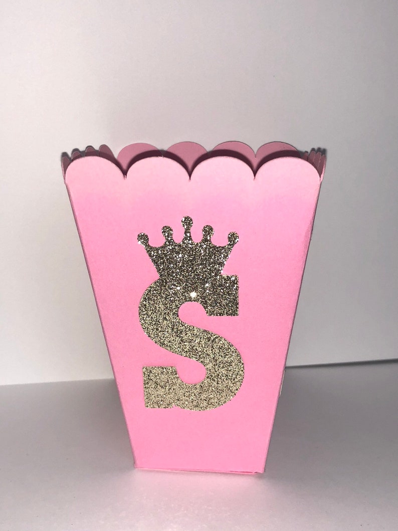 May include: Pink favor box with a scalloped edge. The box has a gold glitter crown and the letter "S" on the front.