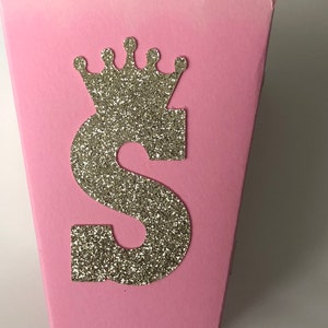 May include: Pink cardboard favor box with a scalloped edge. The box has a gold glitter letter "S" and a gold glitter crown on the front.