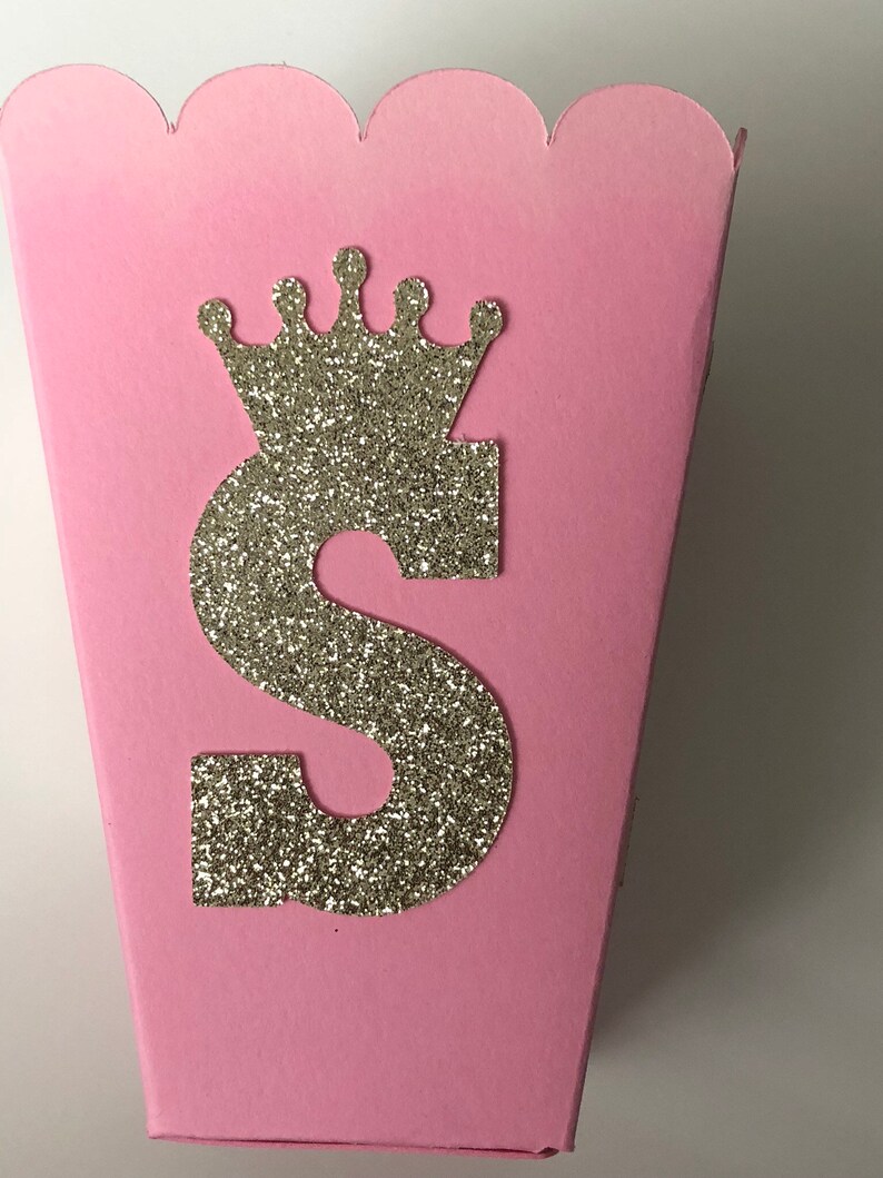 May include: Pink cardboard favor box with a scalloped edge. The box has a gold glitter letter "S" and a gold glitter crown on the front.