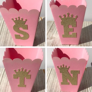 May include: Four pink favor boxes with scalloped edges. Each box has a gold glitter crown and a gold glitter letter. The letters spell out "STEN".