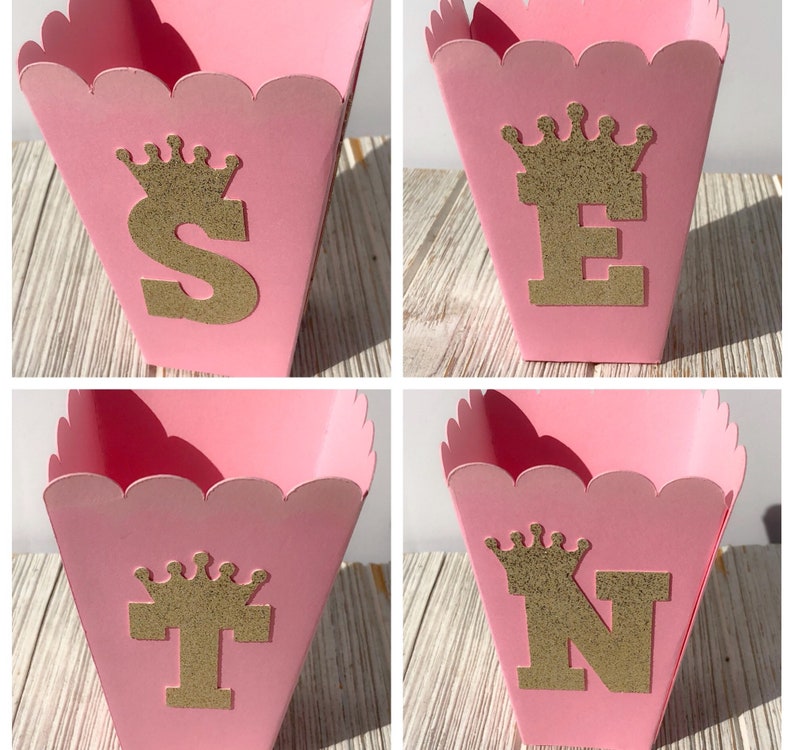 May include: Four pink favor boxes with scalloped edges. Each box has a gold glitter crown and a gold glitter letter. The letters spell out "STEN".