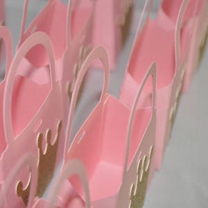 May include: A collection of pink paper gift bags with arched handles. Each bag features a gold glitter crown design on the front. The bags are arranged in rows, suggesting a party or event theme.