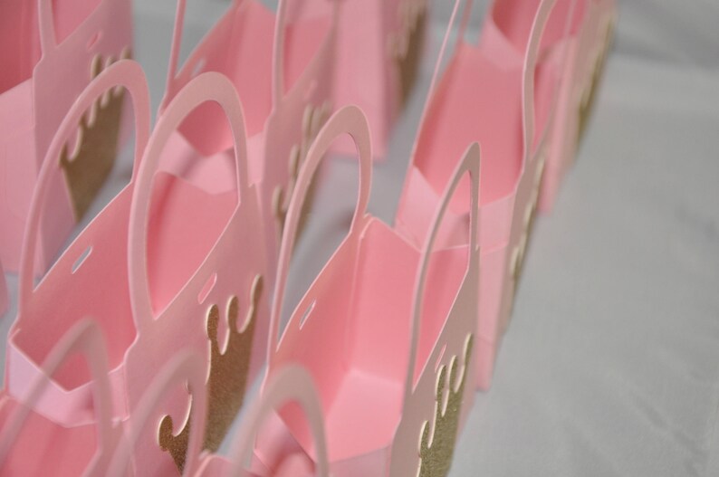 May include: A collection of pink paper gift bags with arched handles. Each bag features a gold glitter crown design on the front. The bags are arranged in rows, suggesting a party or event theme.