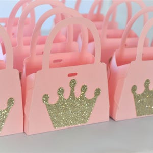 May include: A collection of pink paper purse-shaped gift boxes. Each box features a gold glitter crown design on the front. The boxes have handles and are arranged in rows, suggesting they are for party favors or gifts.