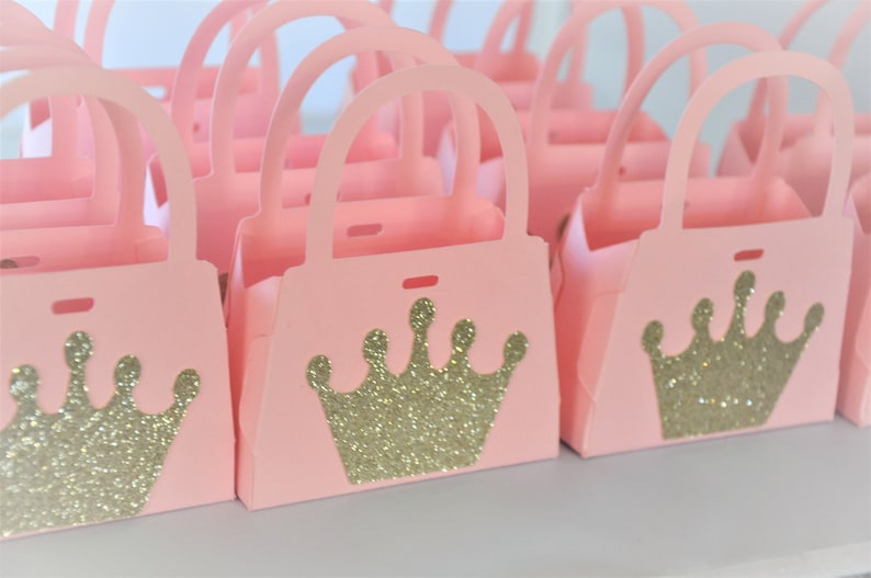 May include: A collection of pink paper purse-shaped gift boxes. Each box features a gold glitter crown design on the front. The boxes have handles and are arranged in rows, suggesting they are for party favors or gifts.