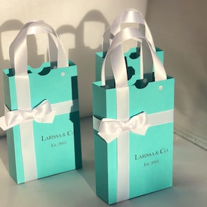 May include: Several small, rectangular gift bags in a light blue color, with white ribbon handles and bows. The bags have a vertical white stripe and the text "LARISSA & Co. Est. 2003" printed on the front.