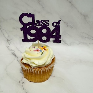 May include: Purple glitter cupcake topper with the text "Class of 1984" on a white cupcake with white frosting and colourful sprinkles.