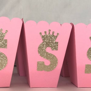 May include: Pink favor boxes with a gold glitter crown and the letter "S" on the front. The boxes are perfect for a princess-themed party.