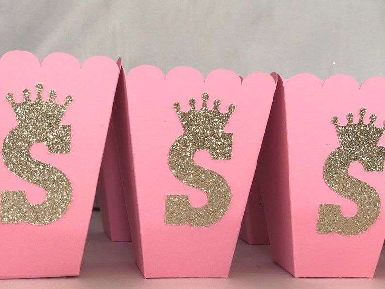 May include: Pink favor boxes with a gold glitter crown and the letter "S" on the front. The boxes are perfect for a princess-themed party.