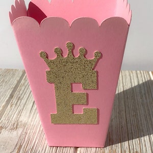 May include: Pink cardboard favor box with a scalloped edge. The box has a gold glitter crown and the letter "E" on the front.
