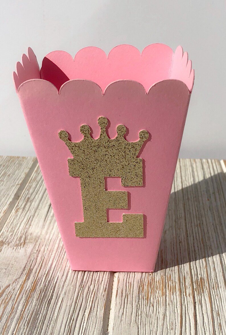 May include: Pink cardboard favor box with a scalloped edge. The box has a gold glitter crown and the letter "E" on the front.