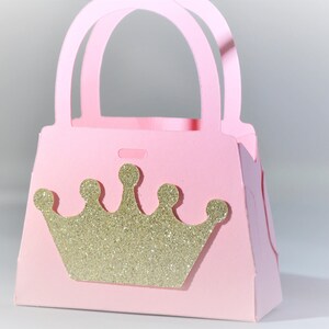 May include: A pink paper purse with a gold glitter crown on the front. The purse has a handle and is designed to hold small items. The overall theme is princess or party favors.