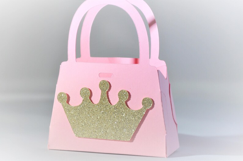 May include: A pink paper purse with a gold glitter crown on the front. The purse has a handle and is designed to hold small items. The overall theme is princess or party favors.