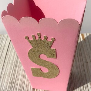 May include: Pink cardboard favor box with a scalloped edge. The box has a gold glitter letter "S" with a gold glitter crown on top.
