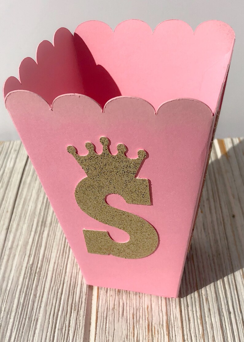 May include: Pink cardboard favor box with a scalloped edge. The box has a gold glitter letter "S" with a gold glitter crown on top.