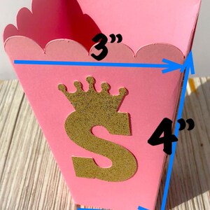 May include: Pink cardboard favor box with a gold glitter letter "S" and a crown on top. The box measures 3 inches wide, 4 inches tall, and 1.75 inches deep.