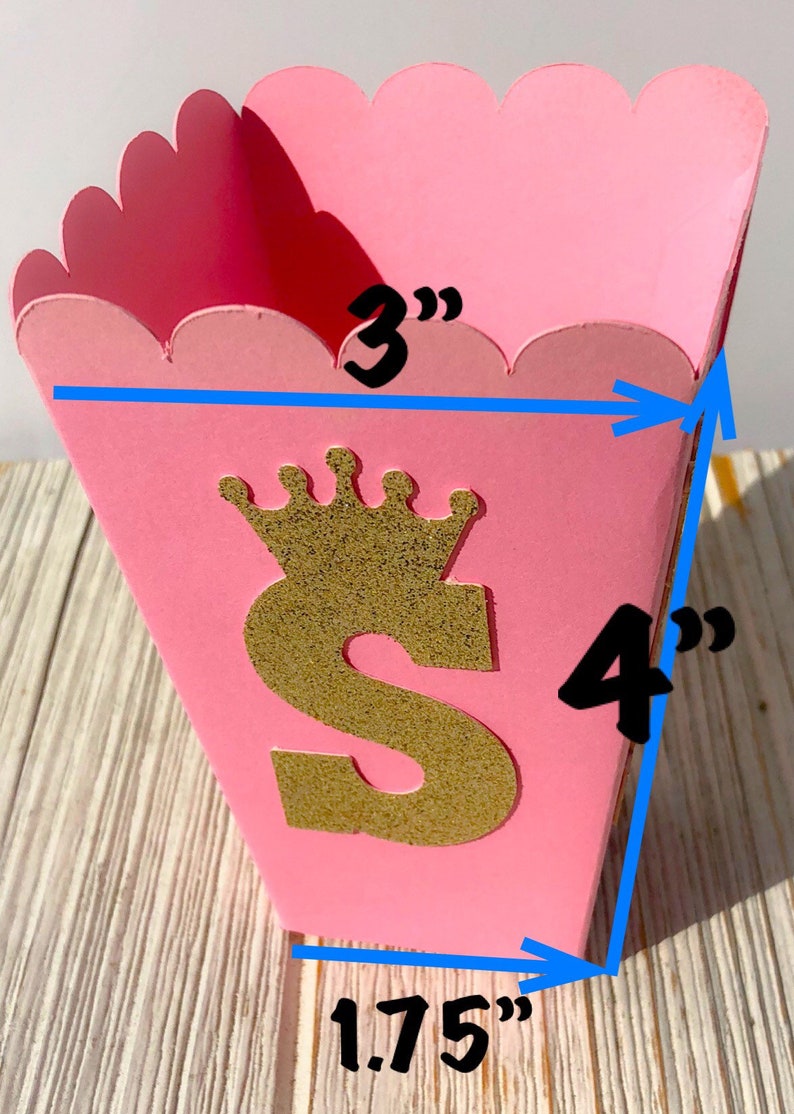 May include: Pink cardboard favor box with a gold glitter letter "S" and a crown on top. The box measures 3 inches wide, 4 inches tall, and 1.75 inches deep.