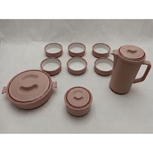 May include: A dusty rose ceramic dinnerware set consisting of a large lidded casserole dish, a smaller lidded bowl, a tall lidded pitcher with a handle, and six small open bowls. Each piece features a matte exterior and a smooth white interior.