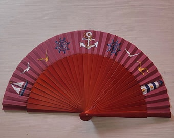 May include: A red wooden hand fan with a nautical theme. The fan features a series of red wooden slats and a decorative top section with images of anchors, sailboats, seagulls, and ship wheels. The fan is approximately 10 inches wide.