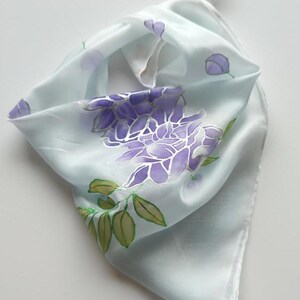 Può includere: Un foulard quadrato azzurro con un disegno dipinto a mano di fiori di glicine viola e foglie verdi. Il tessuto ha una lucentezza liscia e riflettente e il motivo floreale è distribuito sulla superficie con un gruppo centrale.